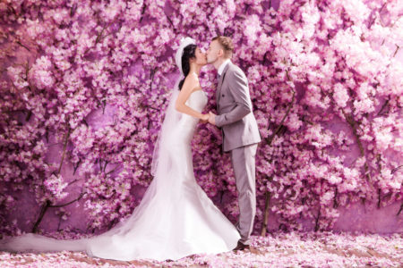 Bride and groom kissing in front of a fun and creative purple flower wall at indoor wedding. 