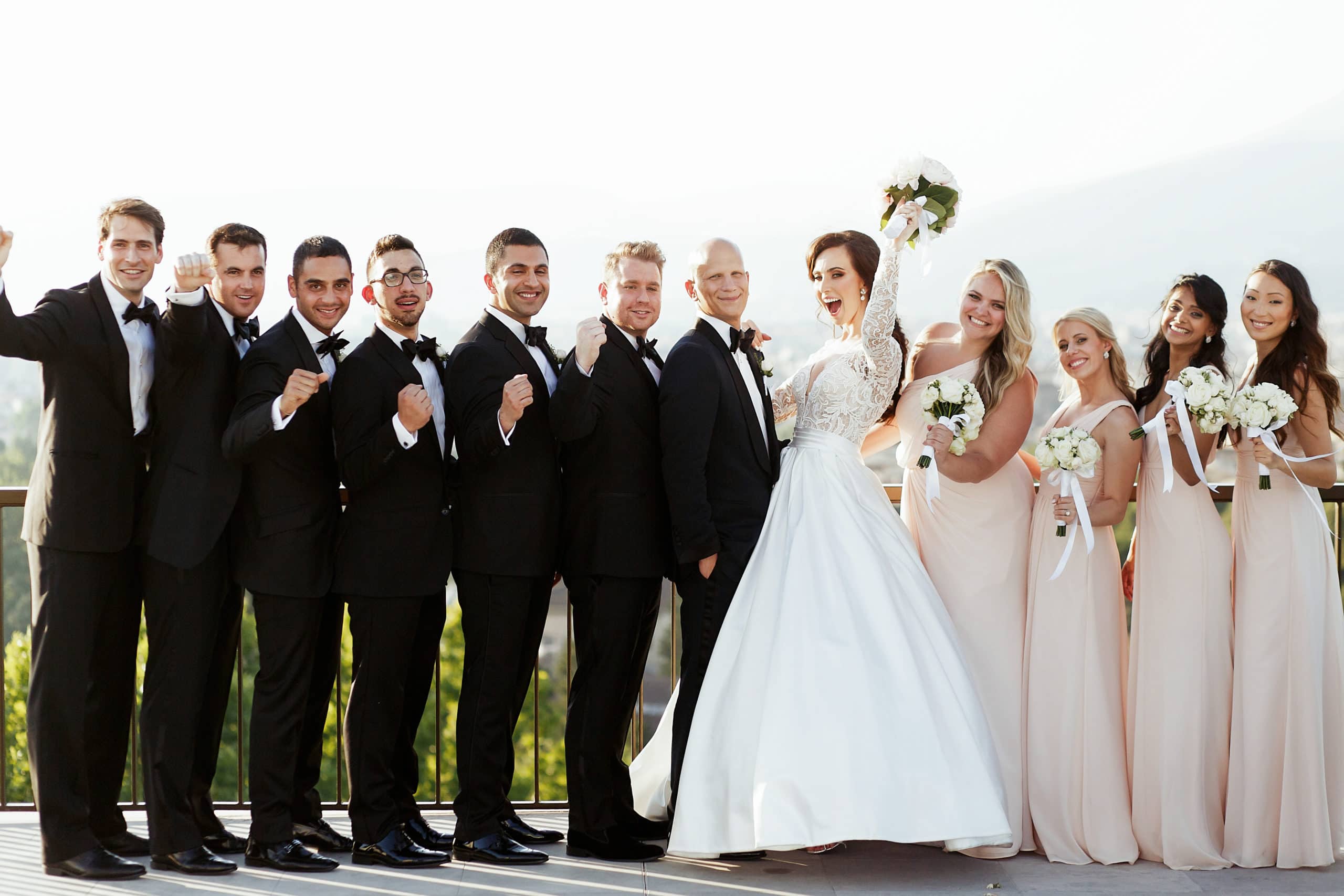 Bridal party in black tuxedos and long blush dresses standing with happy bride and groom. 
