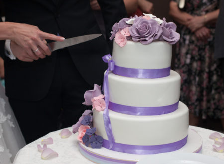 Newlyweds carving delicious white wedding cake with purple roses frosting