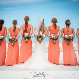 Six bridesmaids and a bride on a Florida beach, holding bouquets facing away from the camera. Bridal bouquet and bridesmaid bouquets by Marco Island Florist.